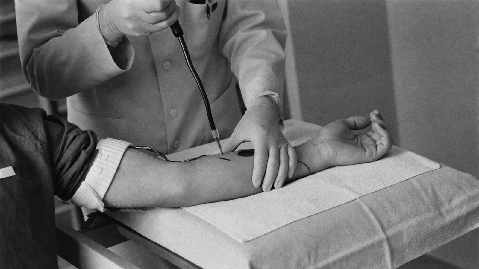 A man's forearm on a clinic table as a gloved phlebotomist prepares a blood draw